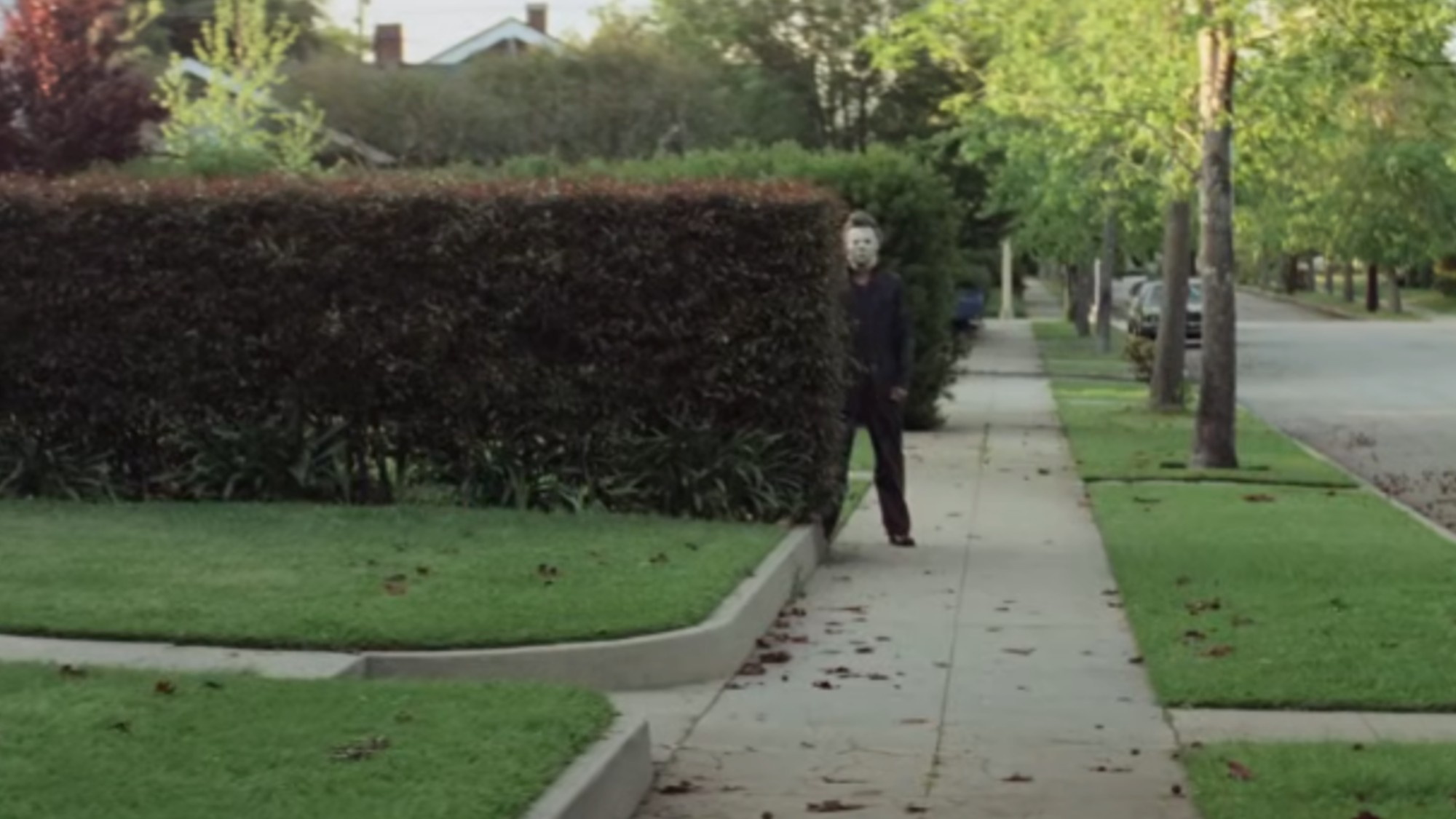 Michael Myers in his trademark white mask and grey coveralls, peering out from behind a hedge on the sidewalk in the first Halloween.