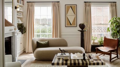 Image of a formal, white living room with two large window with curtains on the back wall. There is a chaise lounge, a striped stone coffee table, a fireplace, and a plant in the corner.