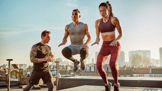 Two people on a city rooftop exercise, jumping and lifting their knees, while a trainer urges them on