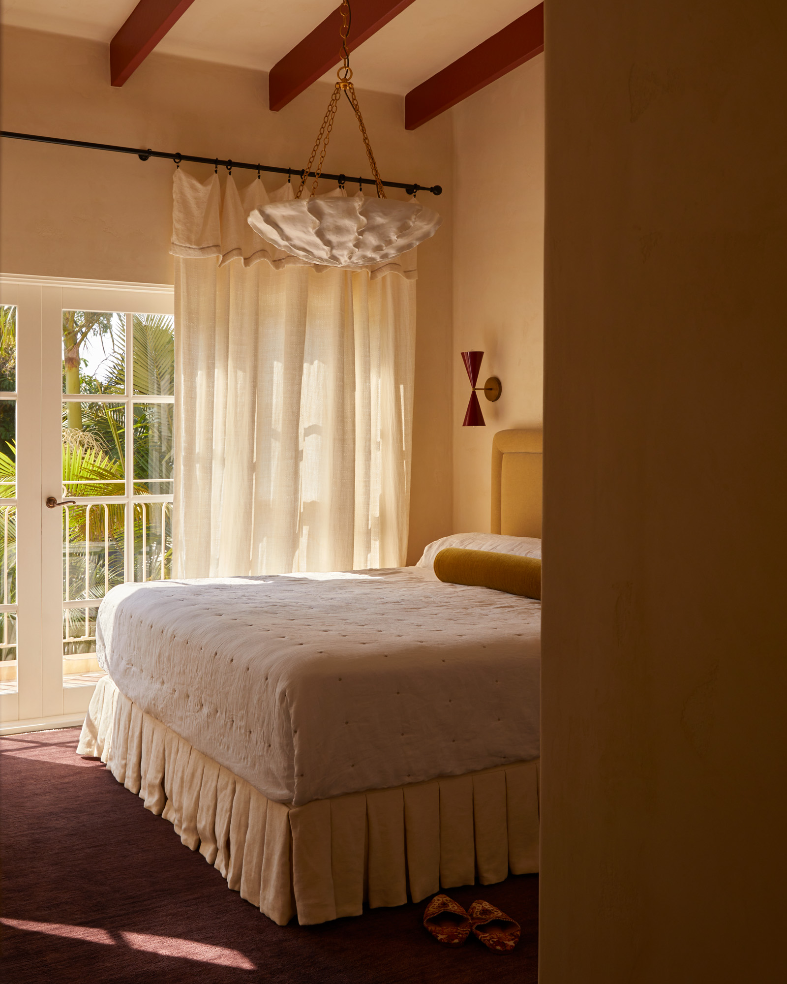 Image of an off white bedroom with french doors that lead to a balcony and have curtains over them. There is a bed with a butter yellow valance and red carpeting in the room.