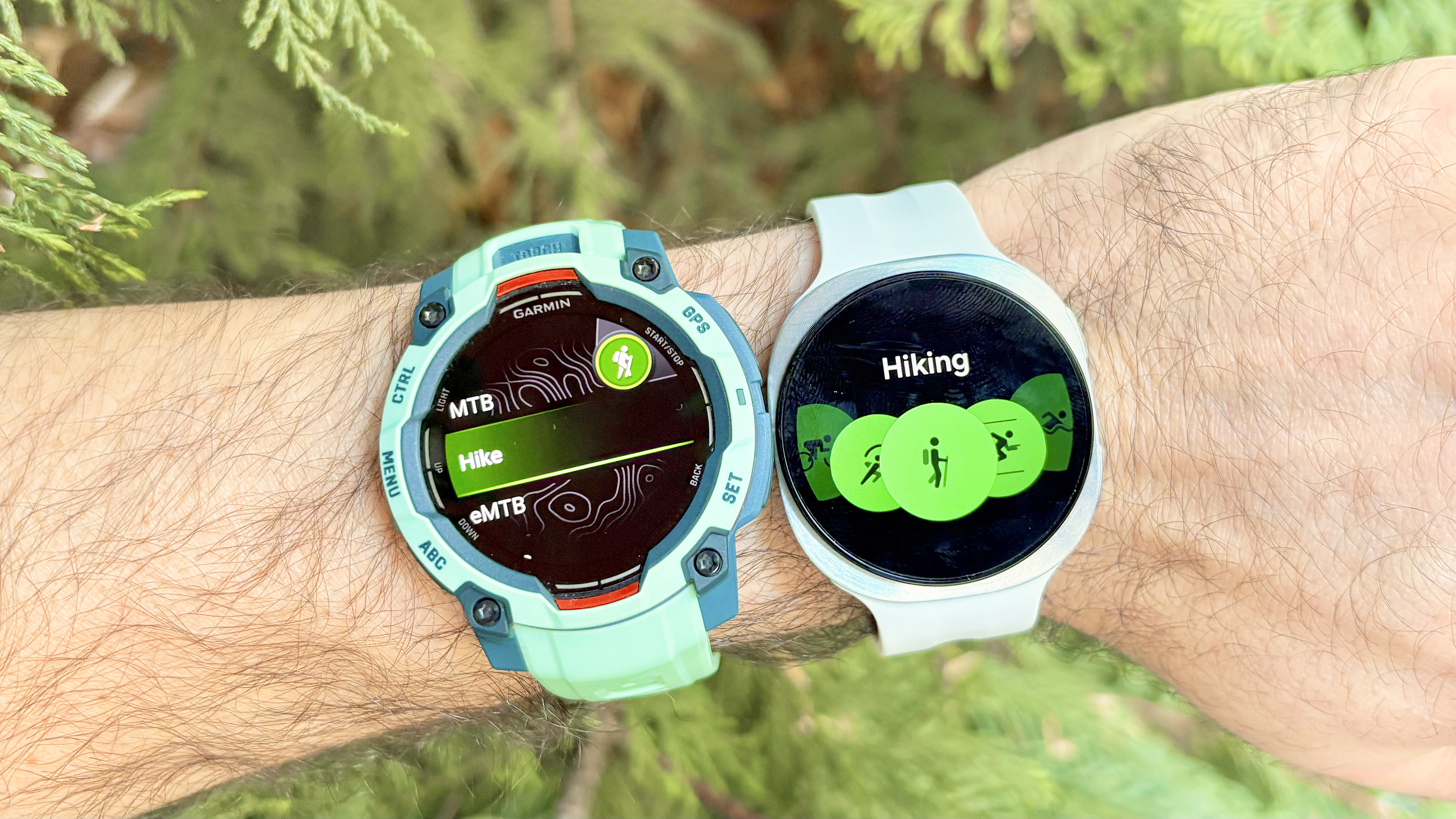 Close-up of the Garmin Instinct 3 next to the Samsung Galaxy Watch 9 with evergreens in the background