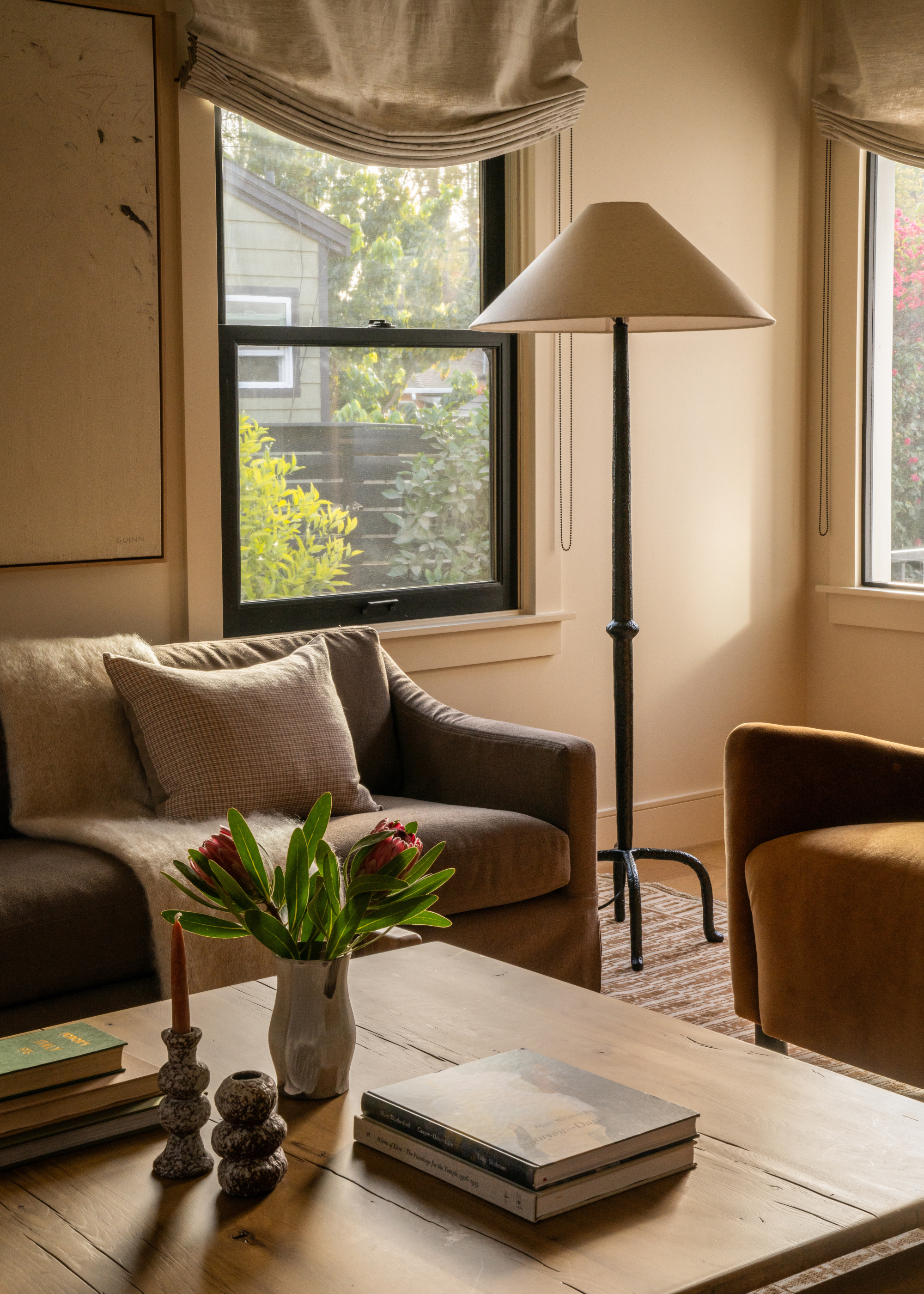 A neutral toned living space with a coffee table styled with books, two candle holders, one with a taper candle, and a small vase of fresh flowers; there is also a tall floor lamp in front of a black framed sash window