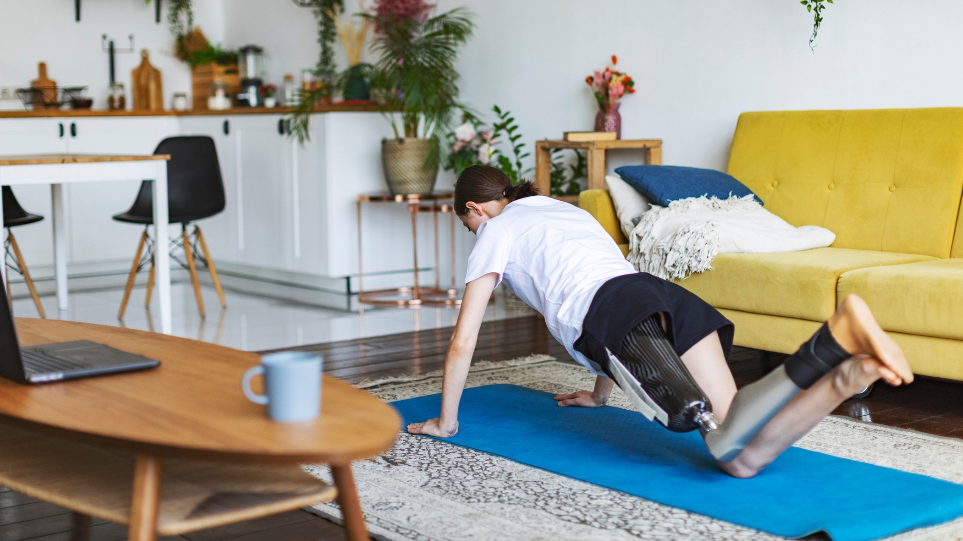 person facing away from the camera with a prothetic leg performing a push up from their knees on a blue exercise mat in a living room setting with a yellow sofa behind them.