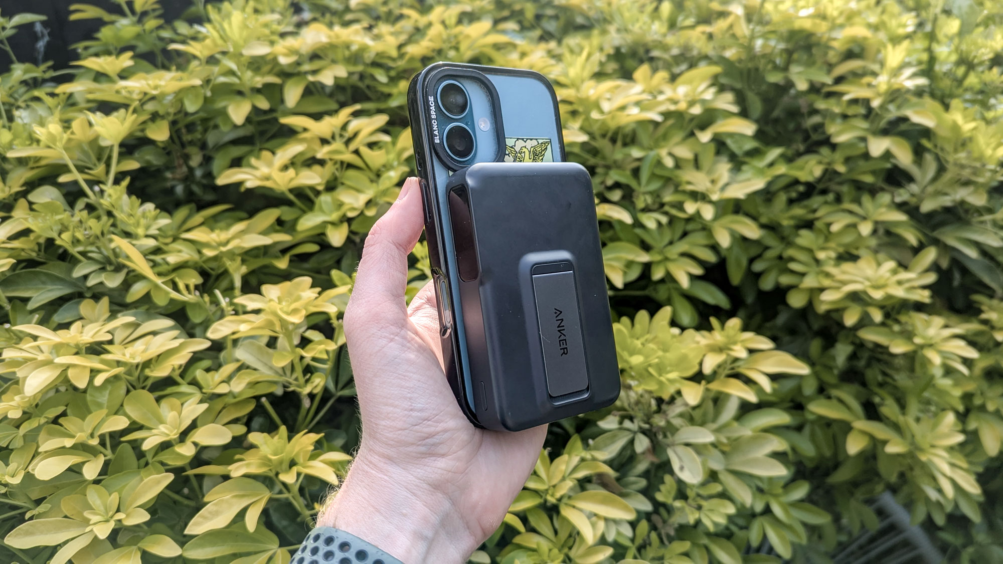 Anker MagGo Power Bank attached to iPhone 16 with case both held in hand with bush in the background