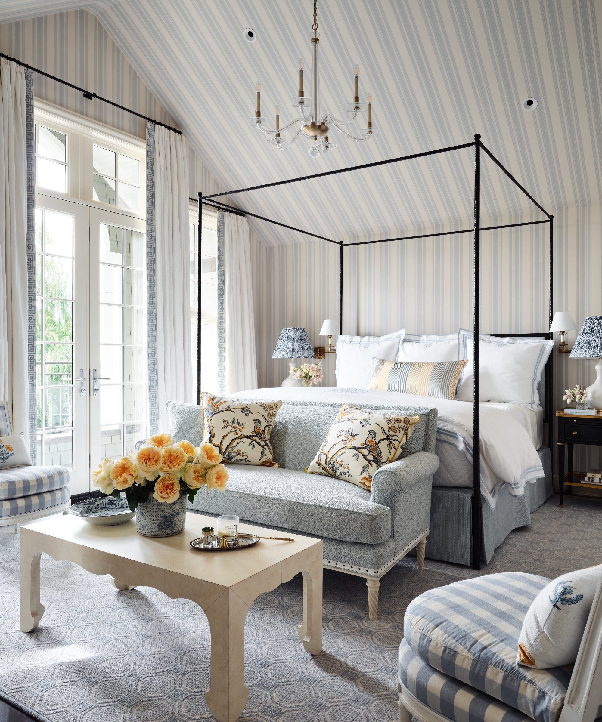 Primary bedroom with all-over blue striped wallpaper, a black four-poster bed, pale blue settee and checked armchairs.