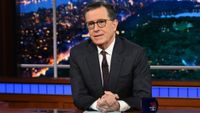Stephen Colbert sits with a blue script and his tea mug at his desk on the Late Show before it wrapped in 2026.