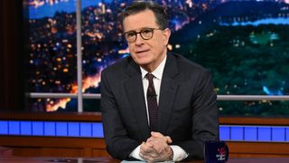 Stephen Colbert sits with a blue script and his tea mug at his desk on the Late Show before it wrapped in 2026.