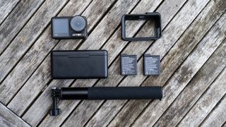 Carefully displayed on a faded wooden table is a small black action camera (DJI Osmo Action 4) and its accessories. From top to bottom, this includes a protective frame, a battery hub/case, two batteries and an extension rod with a mount.
