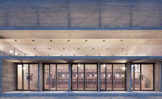 Ground level view of the front doorway of the Library, five black framed glass doors, spot lights shhining onto the entrance, inside wooden foyet and reception desk