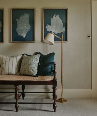 wallpapered hallway with a vintage mahogany bench seat decorated with patterned cushions, a brass floor lamp and a trio of sea themed wall prints