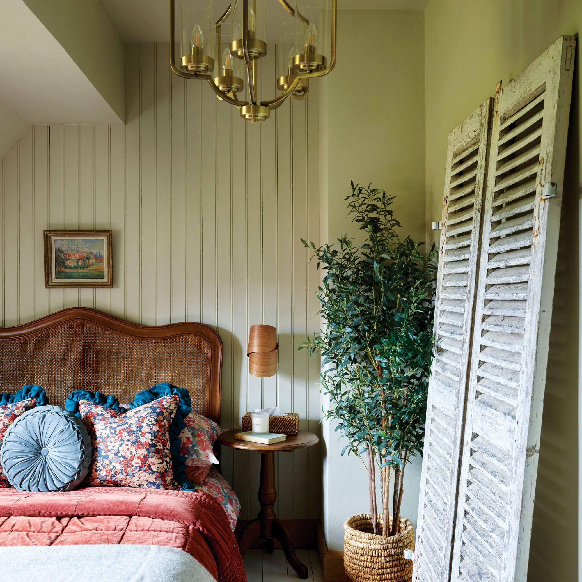 Guest bedroom with green panelled walls, rattan bed frame, wooden bedside table, plant and shutters