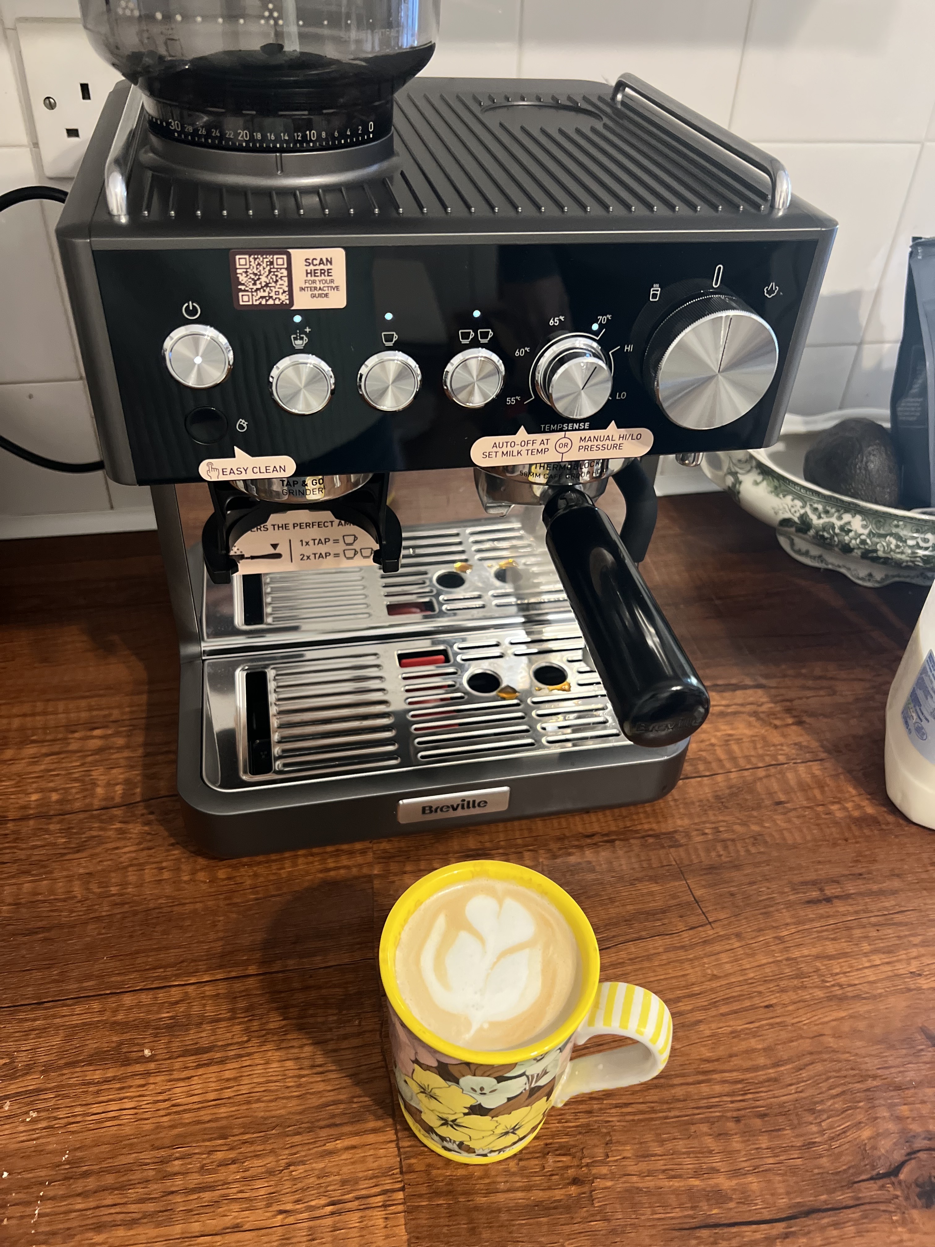 Image of the Breville Barista Sense Espresso Machine in dark gray and black on a wooden countertop with a yellow, floral coffee mug in front of it with a latte that has latte art.