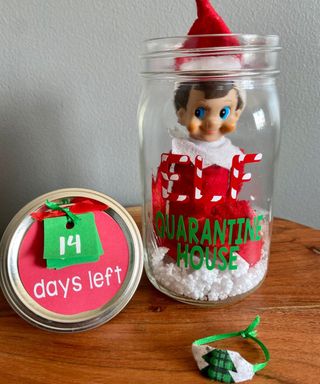 An Elf on the Shelf figurine inside glass mason jar with polystyrene 'snow' decor and decals depicting quarantine house with countdown on lid