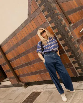 Influencer wears short-sleeve rugby shirt, blue jeans, and white sneakers while leaning against wall.