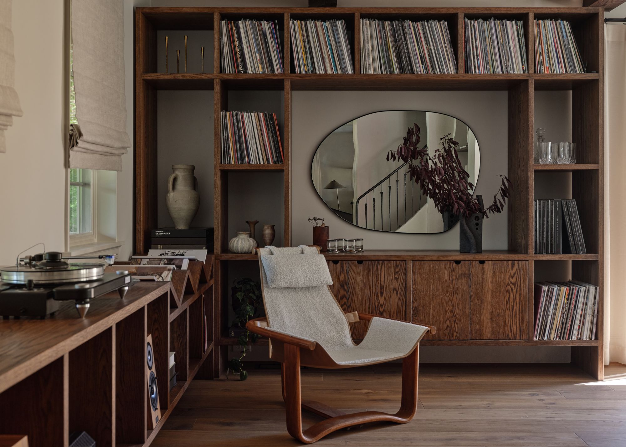 Moody library room with wooden built-in shelving, an accent chair, and a sculptural mirror on the wall