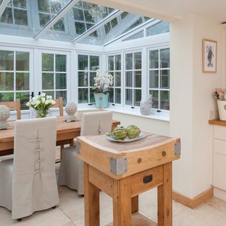 an open conservatory extension just off the kitchen, with glass windows and ceiling with white framing, large wooden dining table and chairs with cream chair covers, and a small wooden island table separating the rooms