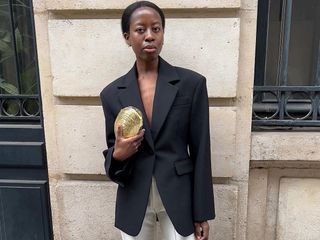 fashion influencer stand on the streets of Paris in a black blazer and metallic gold clutch bag