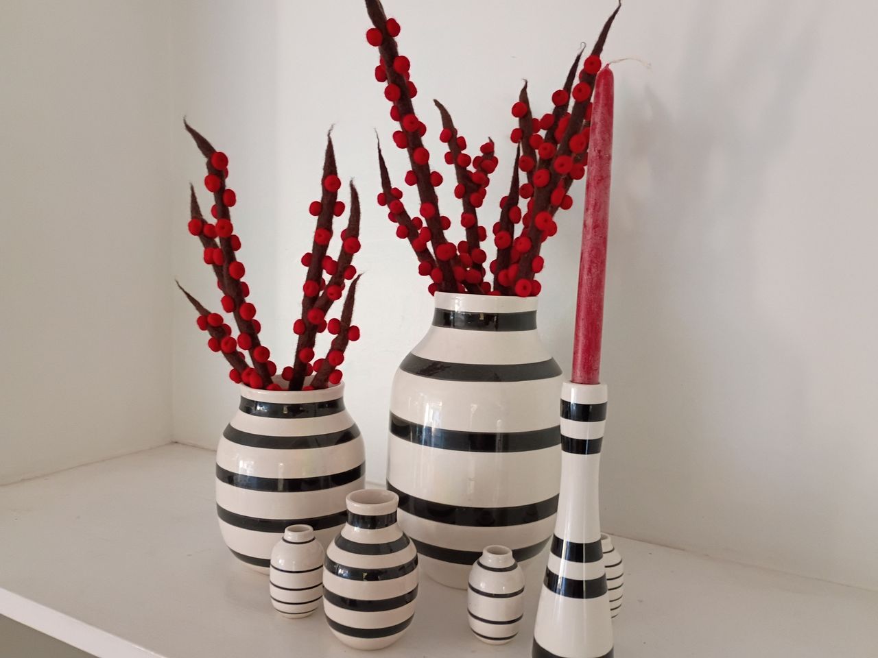 six white vases with black stripes of varying sizes on a white table, two with fake flowers in them and one holding a red candle