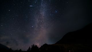 A meteor is pictured streaking the a starry night sky above a silhouetted horizon. The bright band of the Milky Way can be seen tumbling vertically through the centre of the image towards the horizon.