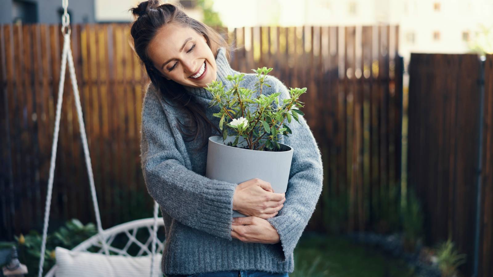 A smiling woman in a sweater holds a houseplant in her backyard