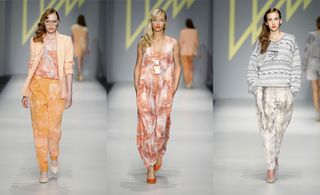 Three models walking on a catwalk. Left: wearing an orange outfit with an orange blazer. Middle: An orange and white print maxi dress. Right: A blue/grey stripe jumper and white/grey trousers.