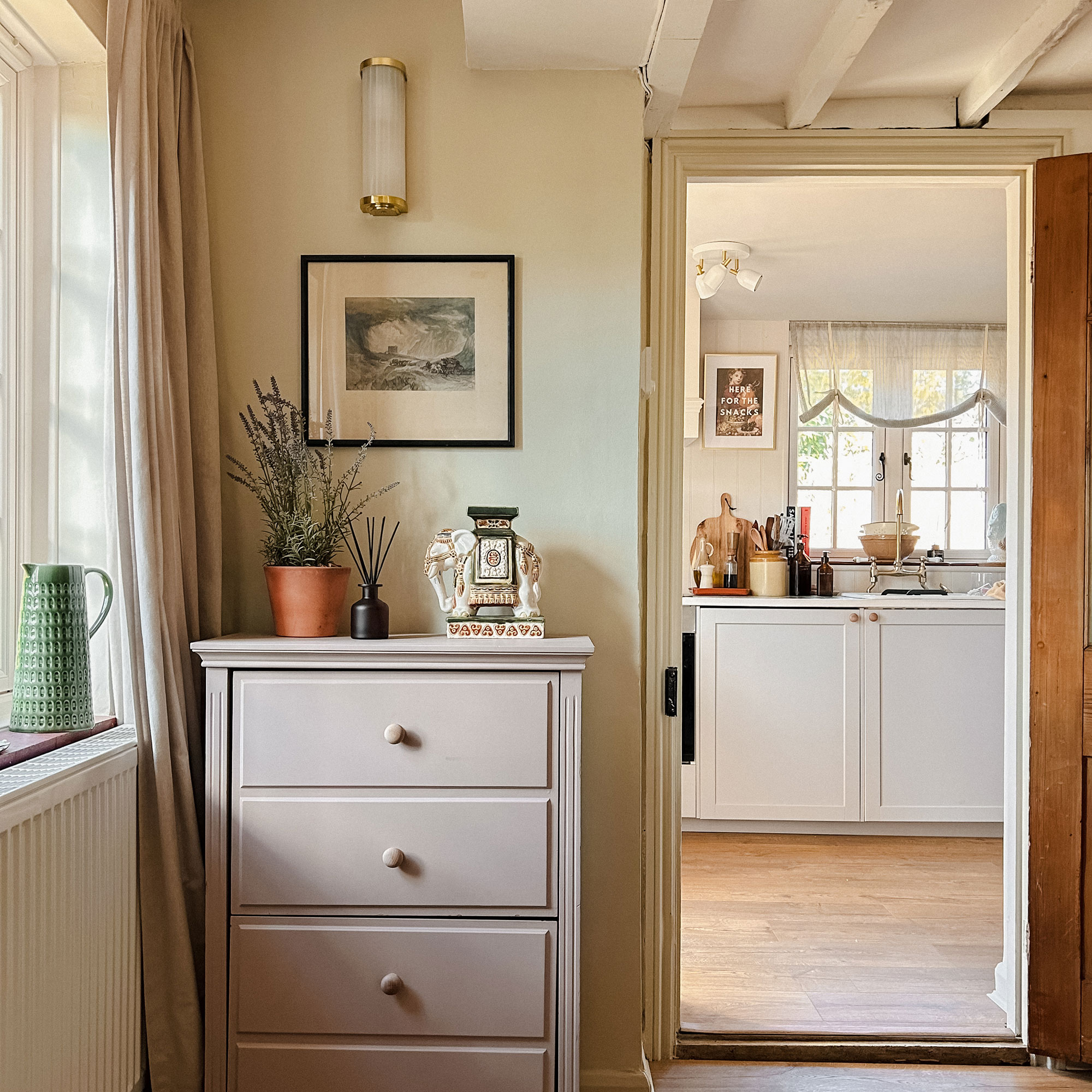 Corner of room with plant and diffuser sitting on a tall boy beneath a painting and a wall light