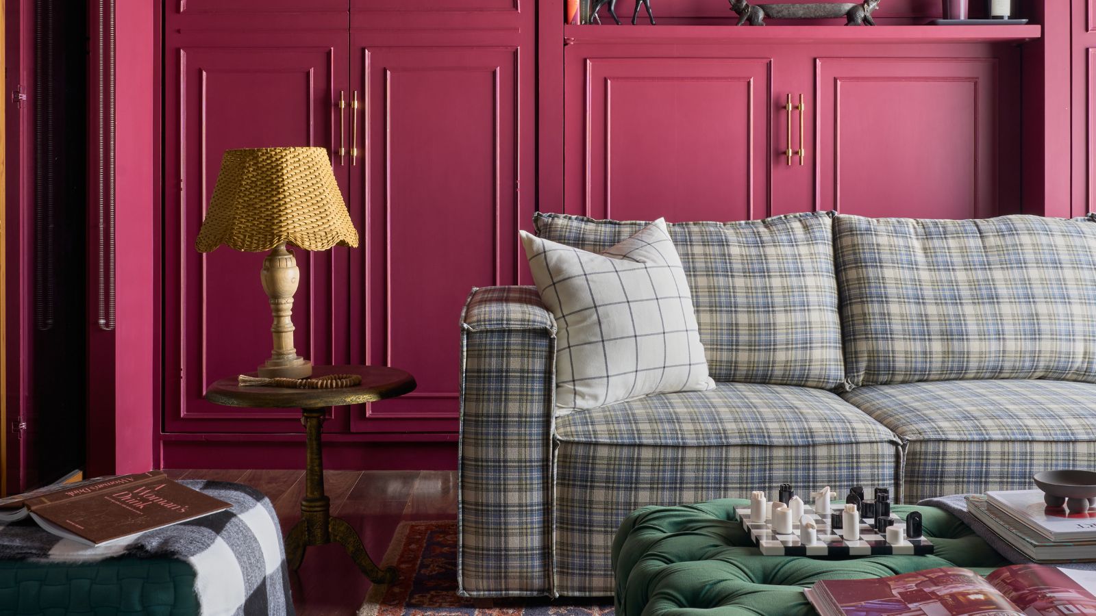 Living room with raspberry pink cabinetry, plaid blue sectional sofa, Persian rug, green velvet ottoman, side table with lamp and plant on the floor