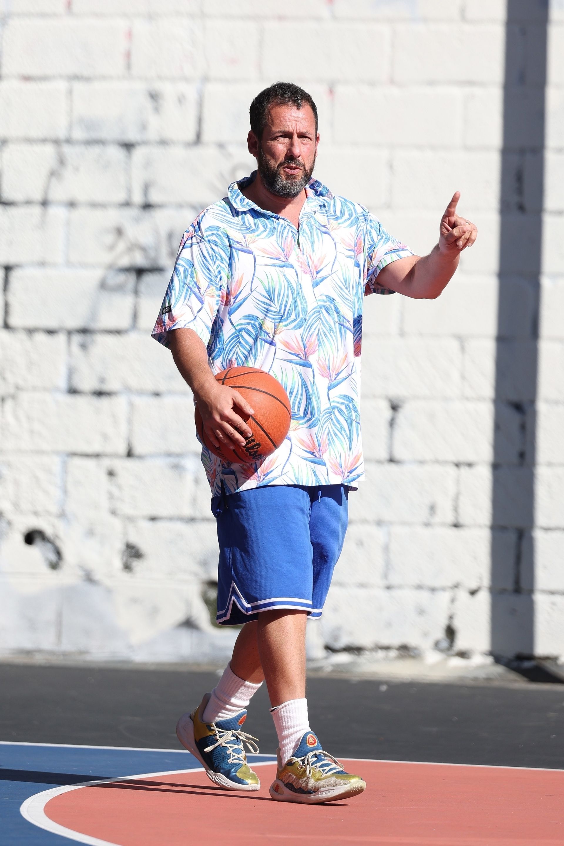 Adam Sandler wearing a patterned polo shirt and blue basketball shorts.
