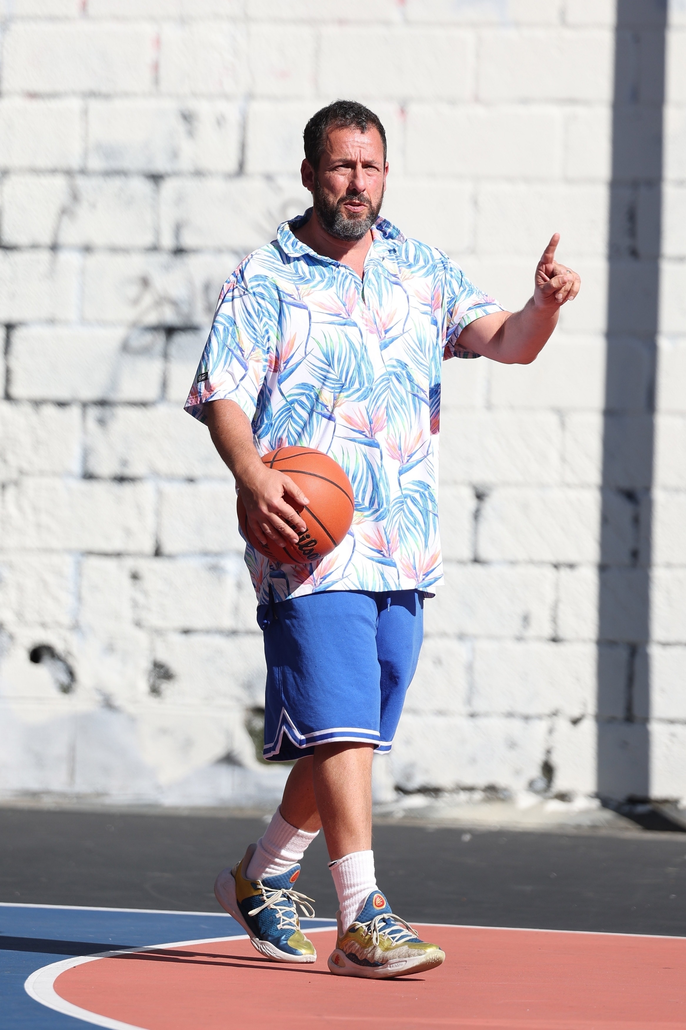 Adam Sandler wearing a patterned polo shirt and blue basketball shorts.