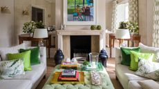 White living room with cozy set up of two sofas facing each other with an upholstered fabric coffee table meets ottoman. Fireplace with large art work above depicting a little cartoon girl playing piano. Green throw cushions and accents layer the room