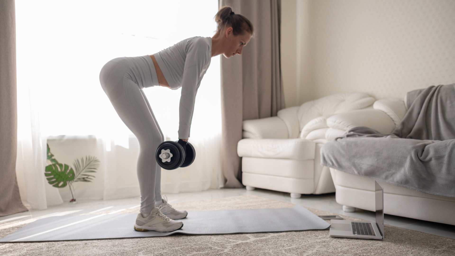 sideways shot of a woman dressed in pale blue top and leggings performing a romanian deadlift in a living room setting with white sofas and a window with long net curtains behind her.