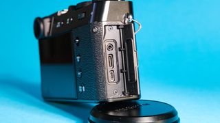 The Fujifilm X100VI mirrorless camera against a blue background propped up using the lens cap with the side ports showing.