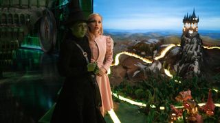 Cynthia Erivo as Elphaba and Ariana Grande as Glinda, standing on the Wicked Part One set next to a miniature of the Yellow Brick Road