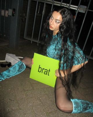 Charli XCX in a blue top and boots holding her green 'brat' album