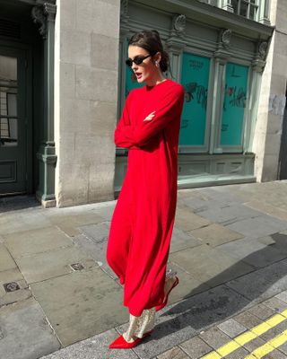 Influencer Valeria Sobalera walks on a London sidewalk wearing black oval sunglasses, a red maxi sweater dress over sequined pants and red pumps