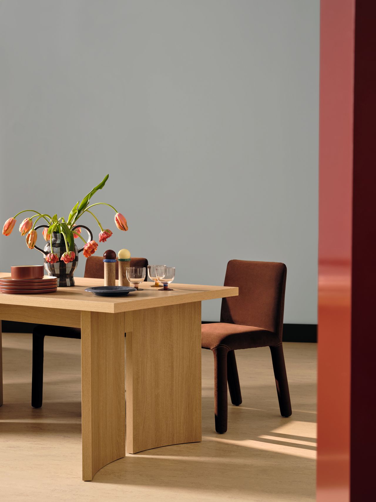 A plywood table in a light blue room with two brown chairs behind it. There is modern tableware on the table and a vase with drooping pink tulips.