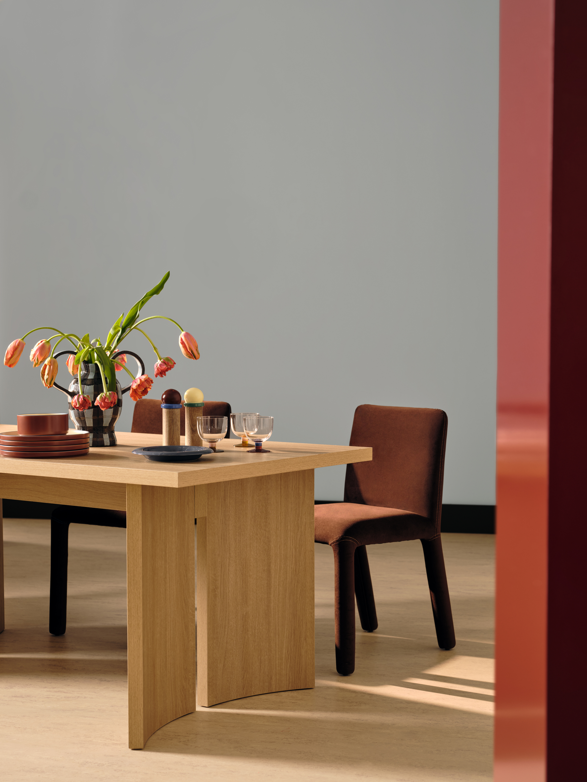 A plywood table in a light blue room with two brown chairs behind it. There is modern tableware on the table and a vase with drooping pink tulips.