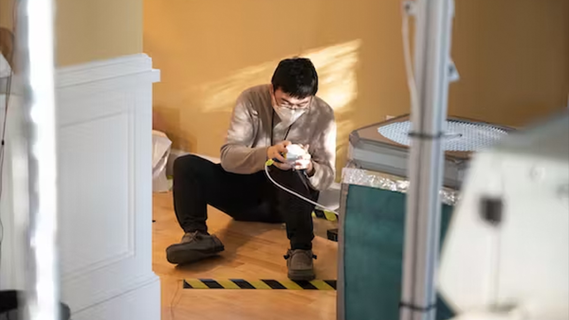 scientist wearing a face mask and glasses sits on the floor of a house while taking samples from a small device in his hands