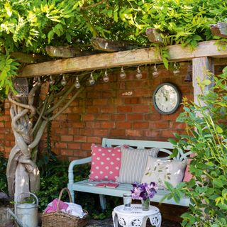 Climbing wisteria over a wooden structure next to an exterior red brick wall with a duck egg blue painted bench with cushions below