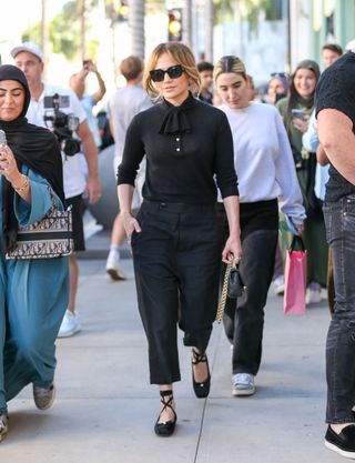 Jennifer Lopez in LA wearing black sunglasses, a black sweater, black cropped trousers, and black lace-up ballet flats.