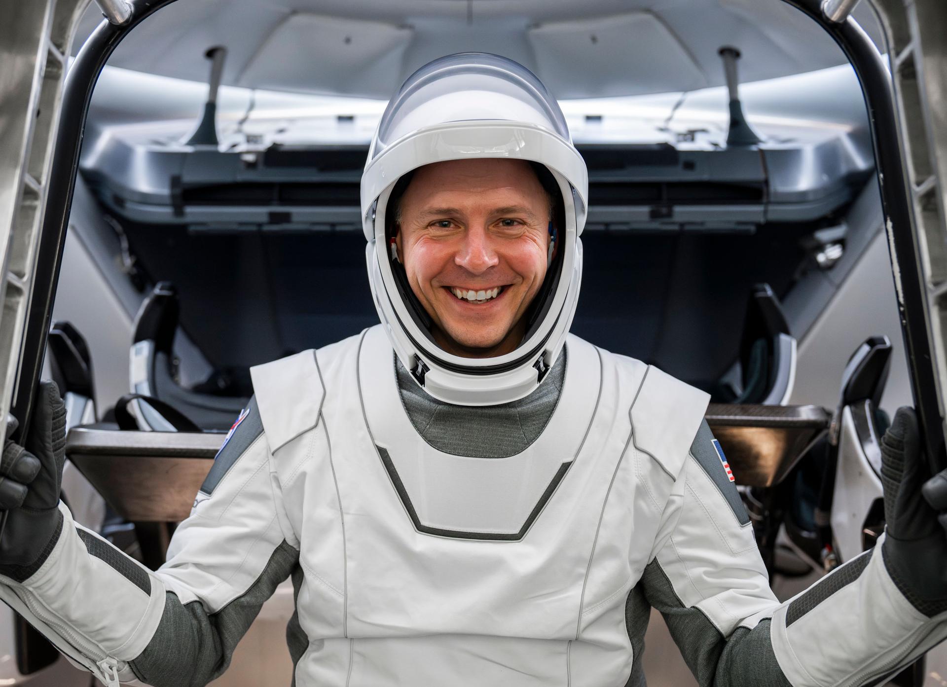 nick hague in a spacesuit smiling and grasping on to the sides of a simulator while standing in front of the camera and smiling