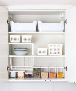 An open white kitchen cabinet with storage bins, storage shelf extenders, and plastic dry goods containers inside of it