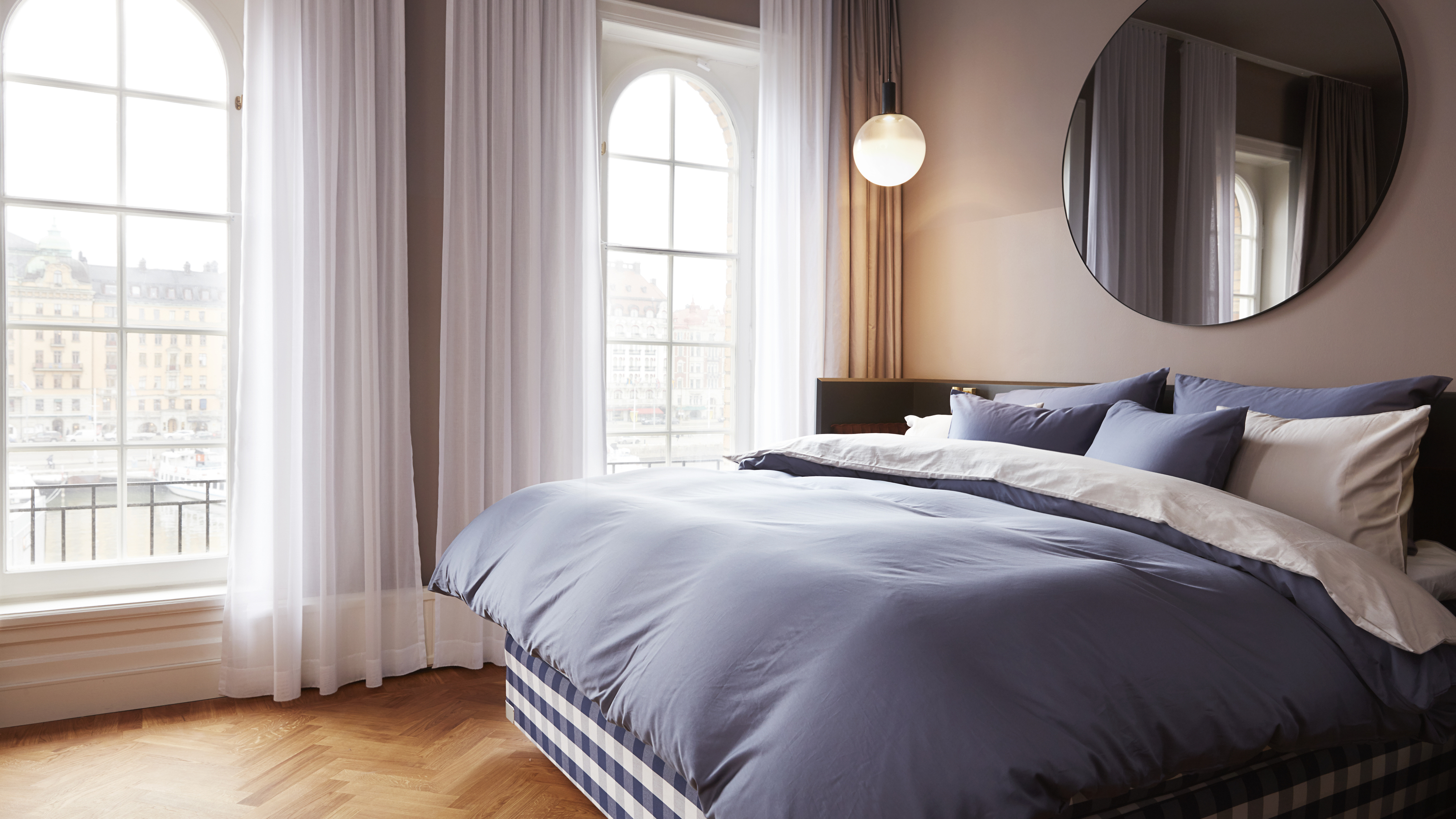 A bedroom with large arched windows, a double bed with blue bedding, and a large circular mirror on the wall