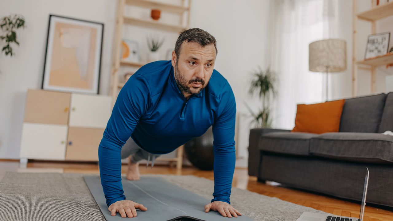 A man in a long-sleeved top performs a push-up at home. Behind him we see a sofa, a chest of drawers, a plant and a lamp.