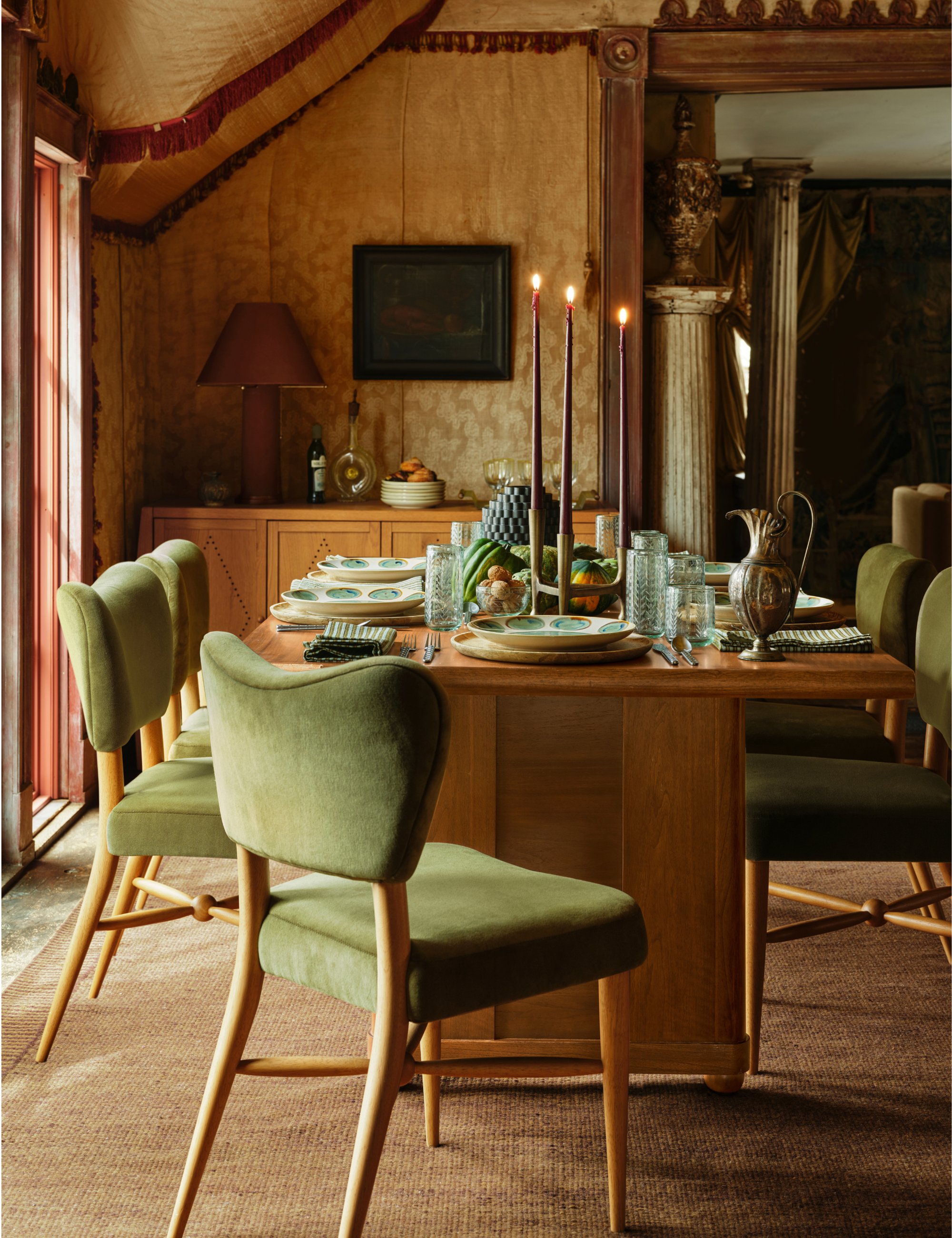 Warm, richly layered dining room featuring green velvet dining chairs, a mid-century-inspired dining table, moiree fabric-covered walls, and dramatically elongated tapers glowing over the scene.