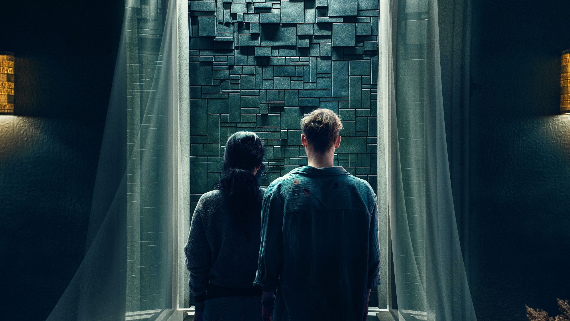 Two people with their back turns looking out a window onto a black brick wall