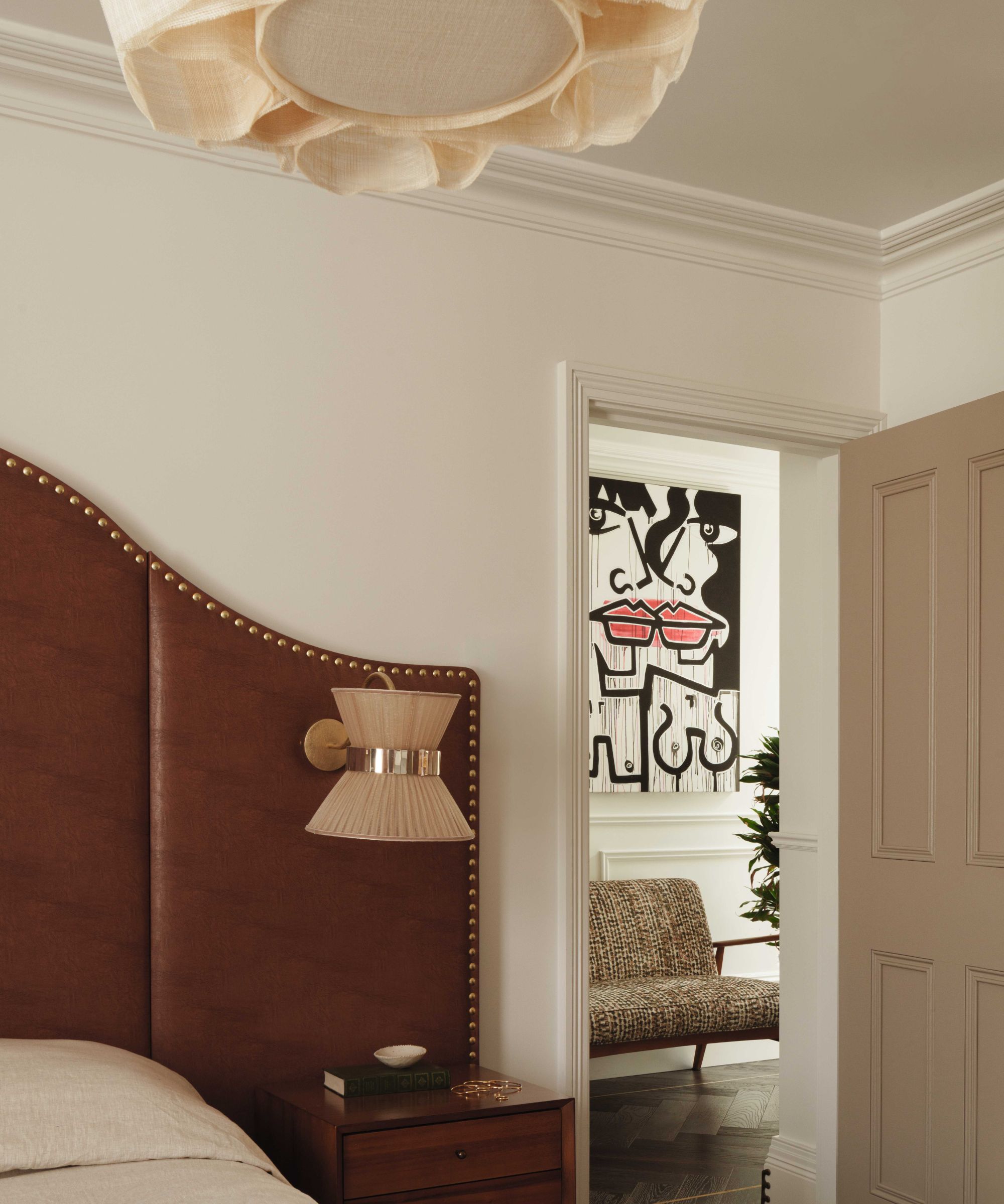 A sophisticated bedroom corner featuring a curved brown leather headboard with brass studding, a walnut nightstand, and a pleated fabric sconce. Through the doorway, a glimpse of a second room reveals a patterned sofa and bold, graphic wall art