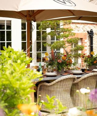 An outdoor dining space with a brown umbrella, a gray table with wine glasses and white plates and bowls, two rattan chairs underneath it, and a house with white windows and doors and brick framing around them