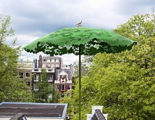 A green parasol made of a leaf pattern with a bird on top of it and buildings and trees behind it.