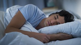 How to fall asleep faster at night: A man with dark hair and wearing a blue t shirt sleeps in a bed dressed with blue sheets